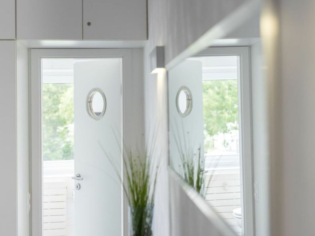 Una puerta blanca con un espejo circular encima que refleja una vista de una cubierta de madera y follaje exterior.