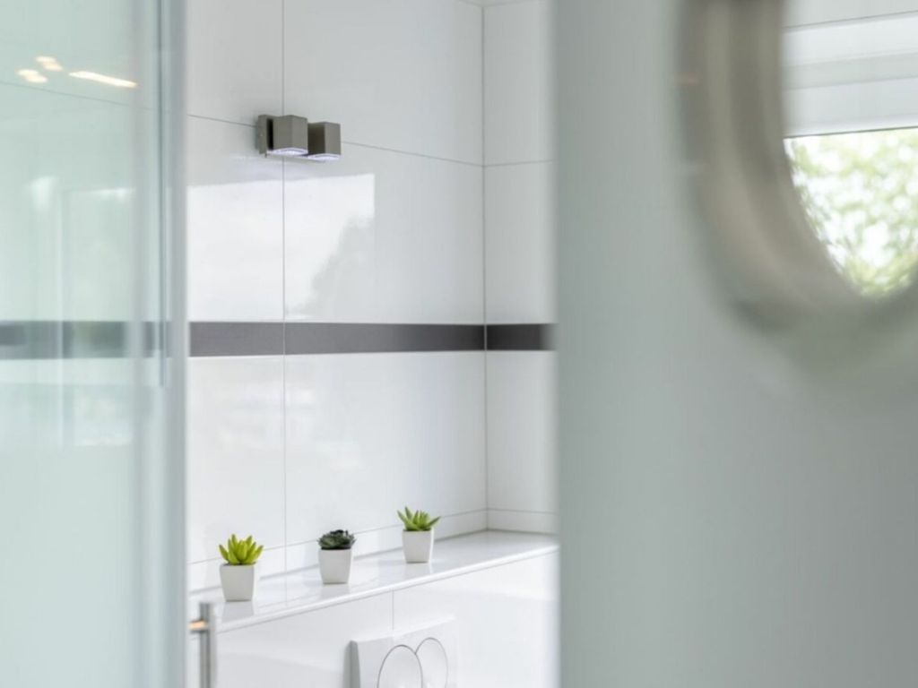 Uma casa de banho moderna com azulejos brancos, cabine de chuveiro em vidro e três pequenas plantas em vasos no balcão.
