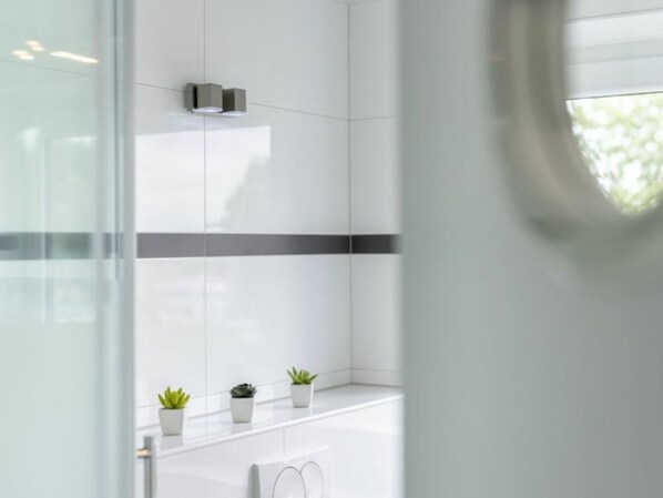 Un baño moderno con azulejos blancos, cabina de ducha de vidrio y tres pequeñas macetas con plantas en la encimera.