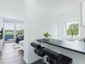 A modern kitchen with a black countertop, two bar stools, a potted plant, and a view of the outdoors through large windows.