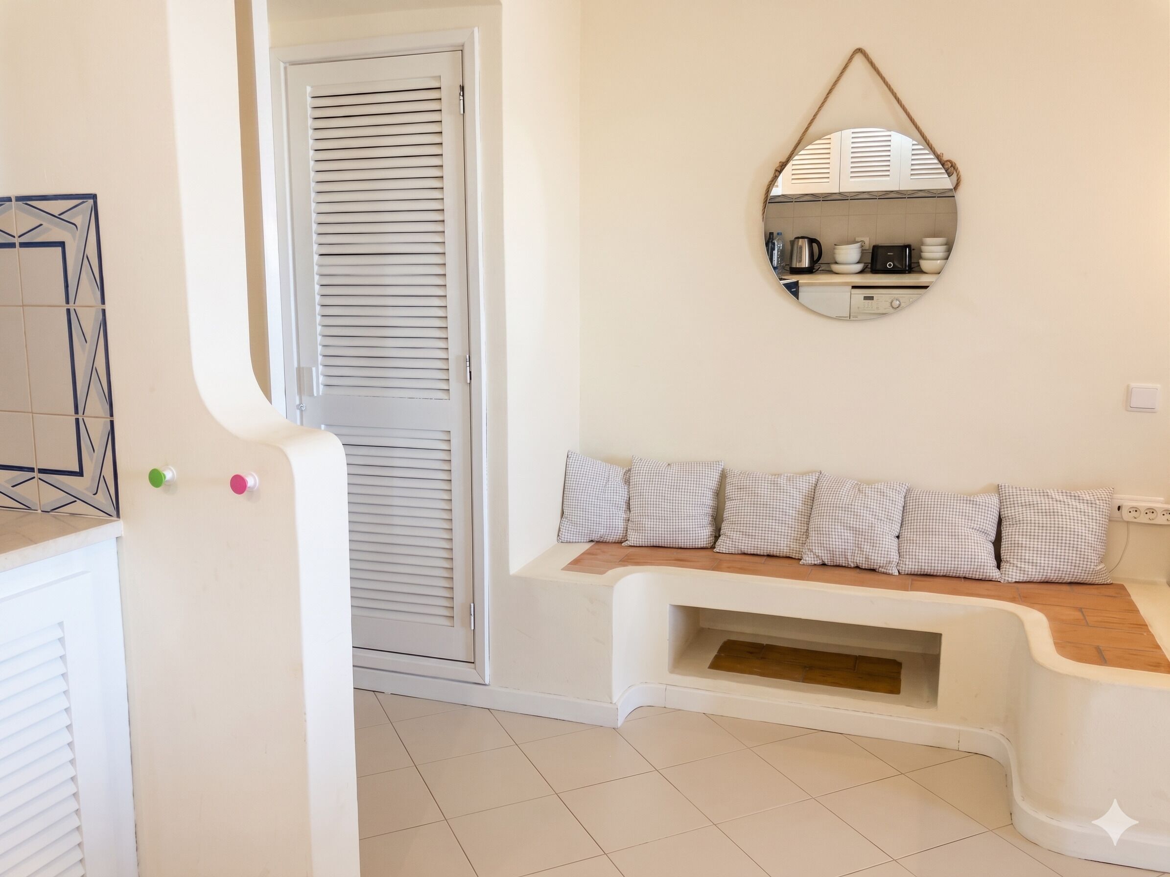 A room with a white door, a bench with checkered cushions, a wall-mounted mirror, and a tiled floor.
