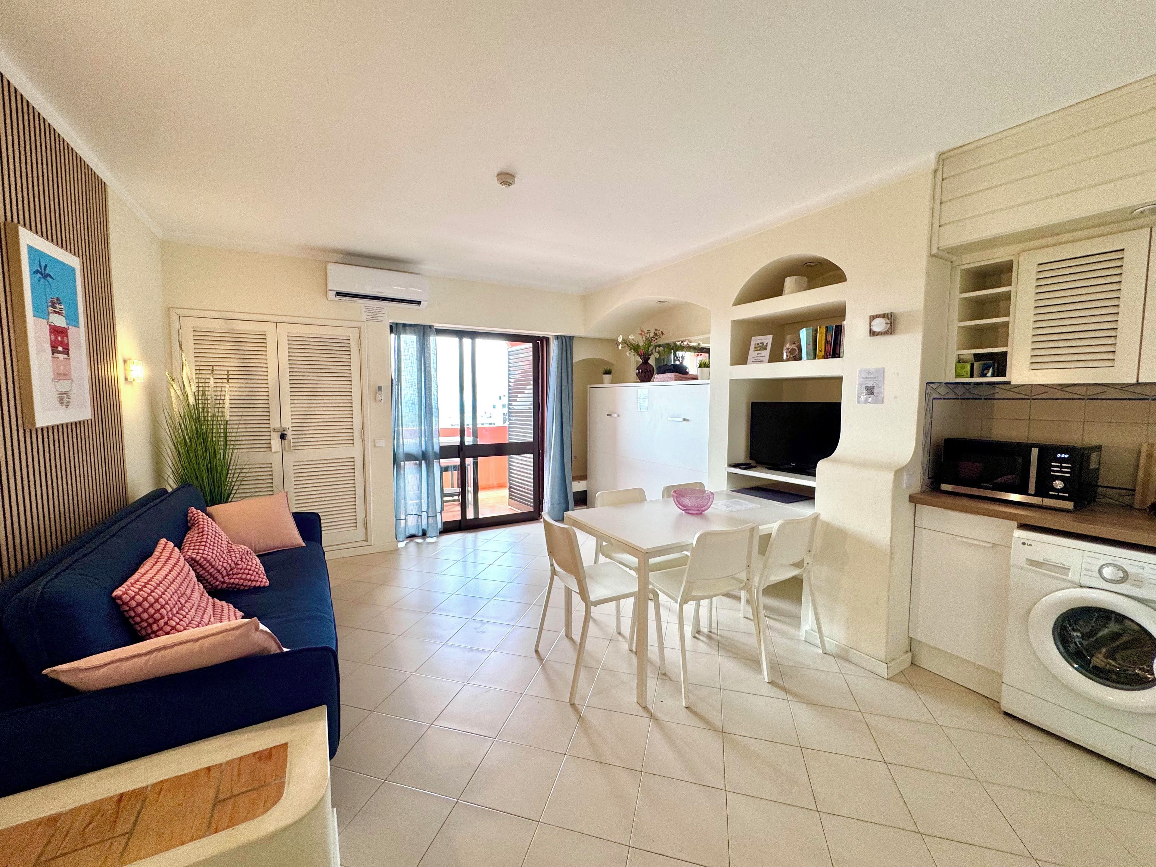 Espaço compacto com sofá, mesa de jantar, cadeiras, cozinha equipada e porta de vidro deslizante que dá para área externa.
