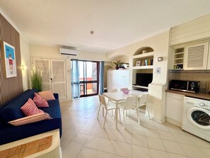 A compact living space with a sofa, dining table, chairs, a kitchen area with appliances, and a sliding glass door leading to an outdoor area.