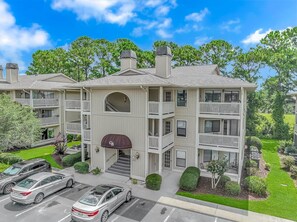 A multi-story building with balconies, a central entrance, and a parking area.