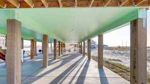 Une allée piétonne couverte, à ciel ouvert, avec des piliers en bois et un toit végétalisé, qui mène à une zone de plage.