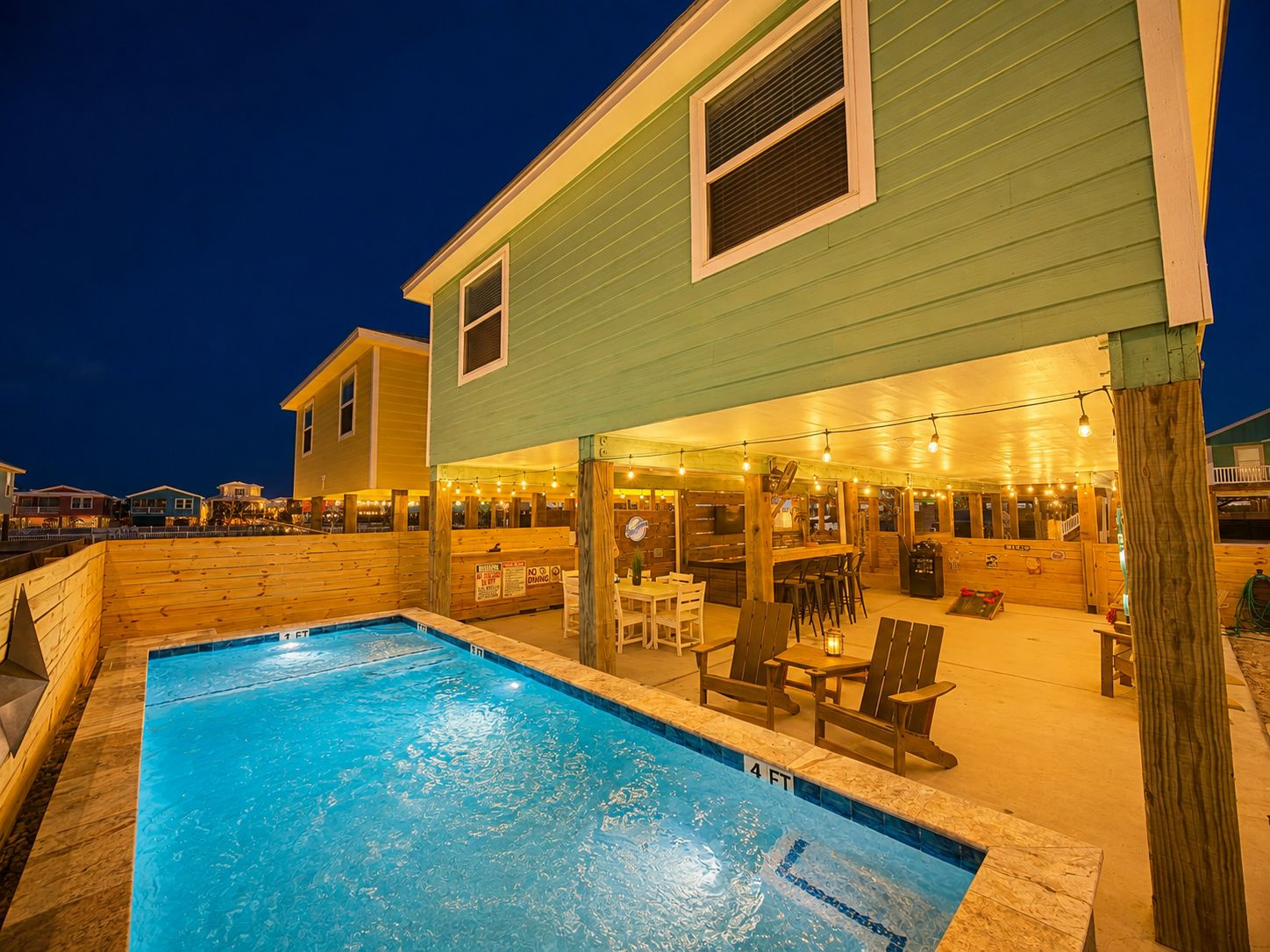 Un espace piscine doté d’un bar et de sièges, entouré d’une terrasse en bois et d’un bâtiment à l’extérieur verdoyant.