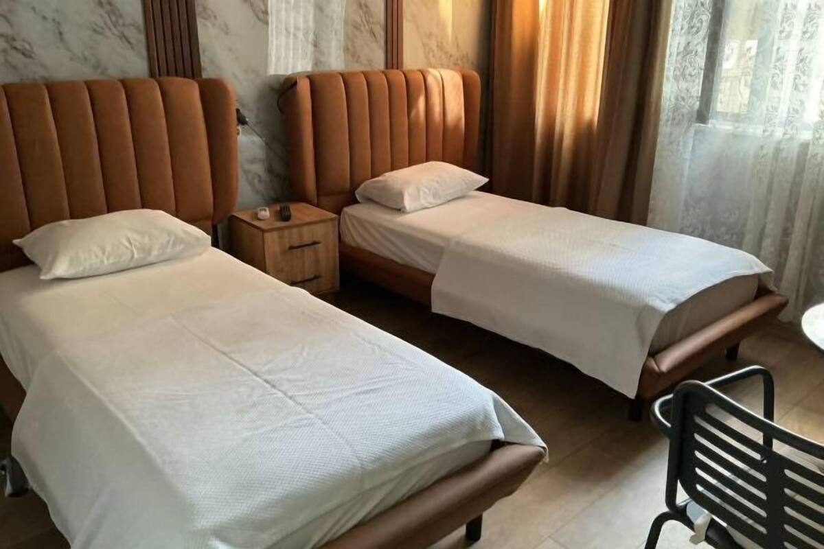 Habitación de hotel con dos camas, una mesita de noche de madera, una pequeña mesa y una silla negra.