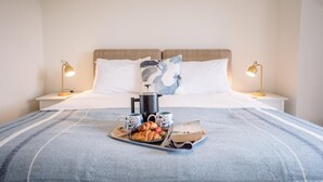 A neatly made bed with a blue and white striped bedspread, two bedside lamps, a coffee maker, a tray with croissants, and a book. - Spindrift, Felixstowe (Felixstowe)