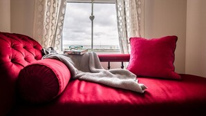 A red sofa with a knitted blanket and a book, a red pillow, and a window with a view of the sky. - Spindrift, Felixstowe (Felixstowe)