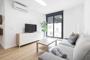 Ein modernes Wohnzimmer mit Fernseher, Couchtisch aus Holz, grauem Sofa und großem Fenster mit Blick auf Gebäude.