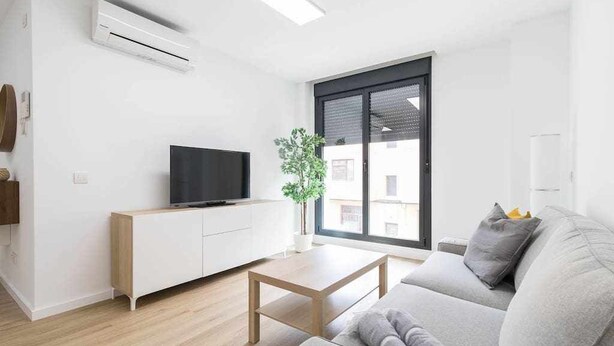 A modern living room with a flat-screen TV on a white cabinet, a grey sofa, a wooden coffee table, and a large window with a view of buildings.