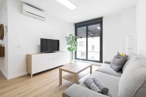 A modern living room with a flat-screen TV on a white cabinet, a grey sofa, a wooden coffee table, and a large window with a view of buildings. - Arrecife Canteras Apartment (Palmas De Gran Canaria, Las)