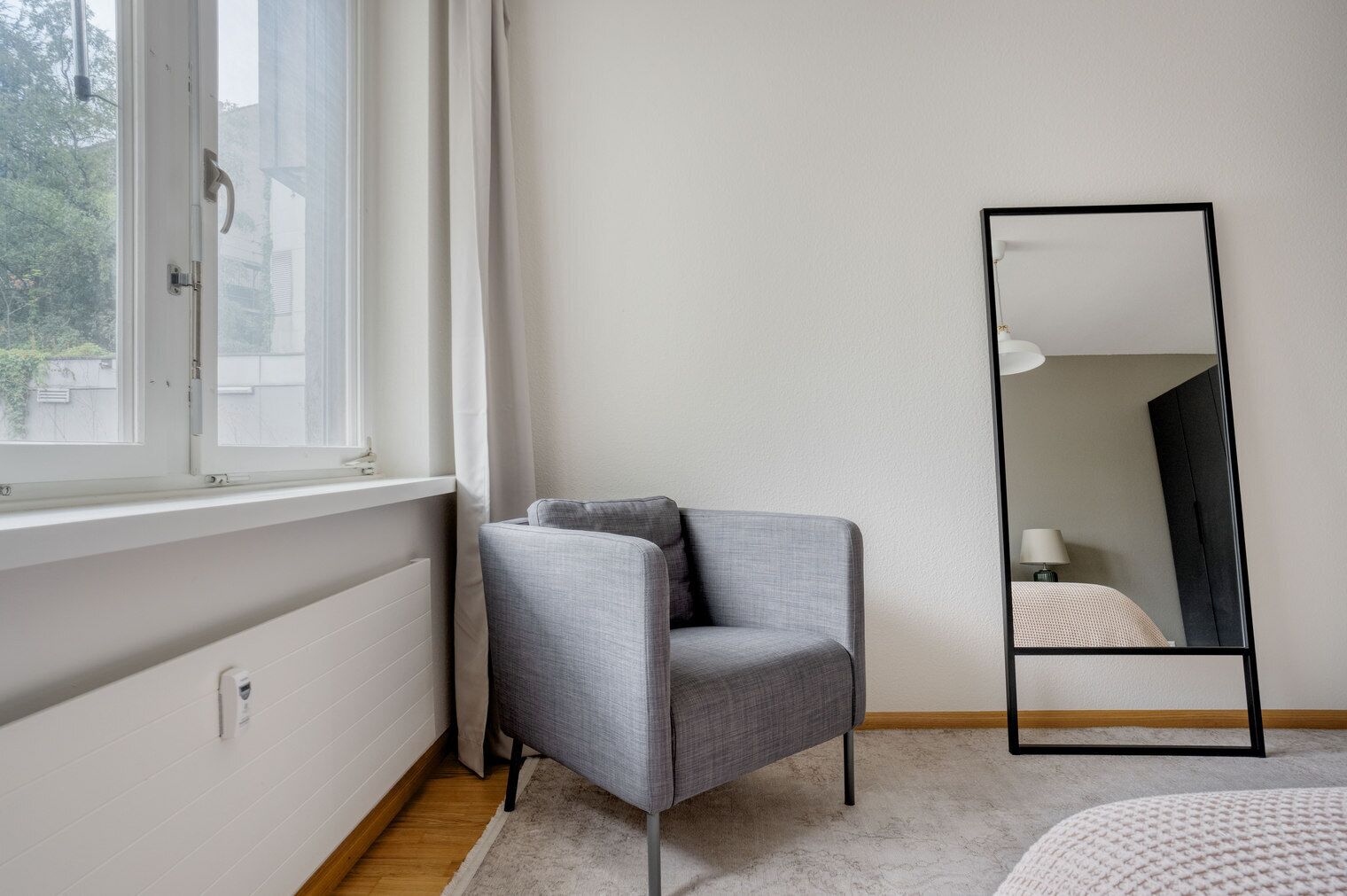 A room with a grey armchair, a large mirror, and a window with curtains.