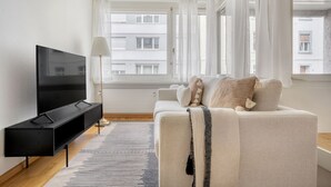 A modern living room with a flat-screen TV, a sofa with cushions, a floor lamp, and a view of buildings through large windows. - Superb Gellert 2 Room w/ Storage Room, nr Tram & Market, by Blueground (Basel)