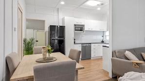 A modern kitchen with white cabinets, a black refrigerator, and a microwave. There is a dining area with a wooden table and chairs, and a grey sofa in the background. - "Suttons Beach Breeze" - Margate Q 4109 (Margate)