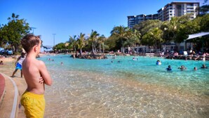 A beach with clear turquoise water, palm trees, and a building in the background. - "Suttons Beach Breeze" - Margate Q 4109 (Margate)