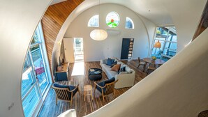 A spacious living area with a high ceiling, wooden flooring, and large windows. There is a staircase leading to an upper level.