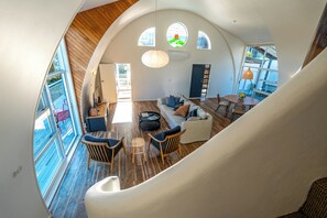 Ampla sala de estar com pé direito alto, piso de madeira e grandes janelas. Escada leva a um andar superior.