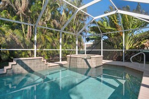 Una piscina con cerramiento de vidrio, rodeada de palmeras y un solárium con baldosas.