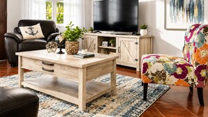 Een woonkamer met een houten salontafel, een zwarte leren bank, een kleurrijke fauteuil, een flatscreen televisie en een schilderij aan de muur.