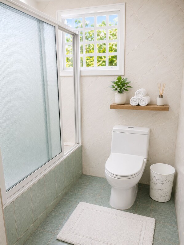 A bathroom with a toilet, a marble trash can, a white rug, a window with a view of greenery, and a shelf with a plant and rolled towels.