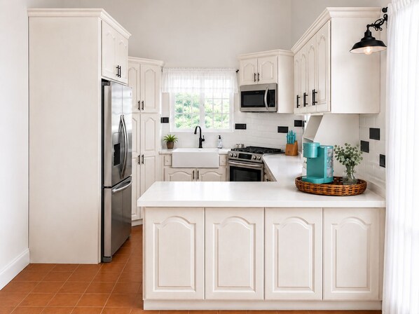 A kitchen with white cabinetry, stainless steel appliances, a window with white curtains, and a teal-colored island with a basket and a vase.