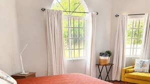 Ein Schlafzimmer mit einem hölzernen Bett, einem Nachttisch mit Lampe, einem kleinen Tisch mit Pflanze und einem gelben Sessel.
