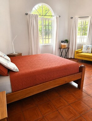 A bedroom with a wooden bed, a bedside table with a lamp, a small table with a plant, and a yellow armchair.