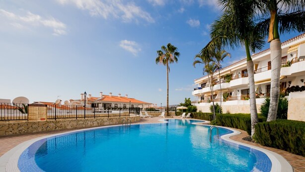 A swimming pool with a tiled edge, surrounded by palm trees and a building with balconies. - 21 C Apartamento Las Bouganvillas (Costa Adeje)
