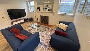 Una sala de estar moderna con un televisor de pantalla plana grande, dos sofás azules con cojines rojos, una mesa de centro de vidrio y una alfombra estampada.