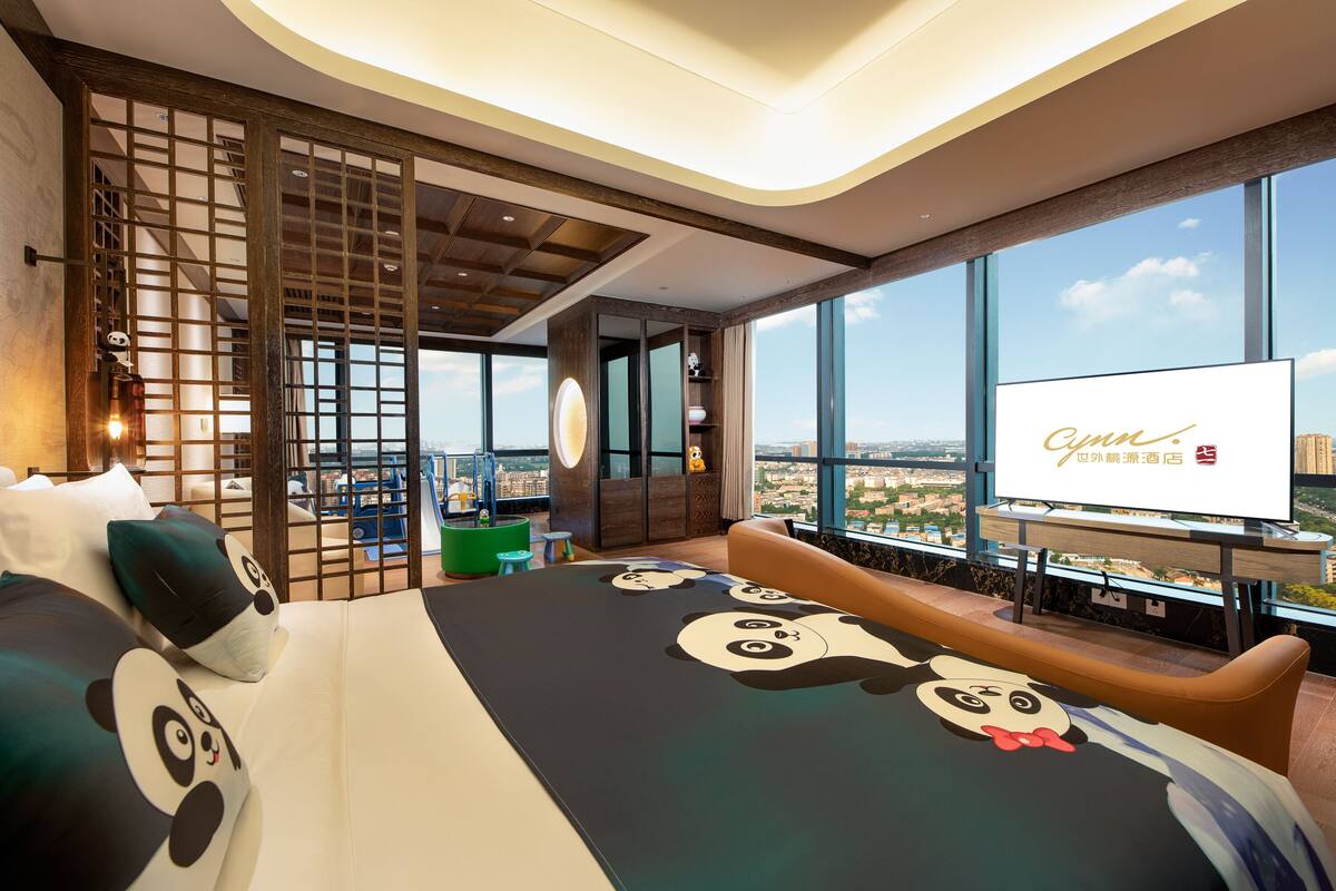 A hotel room with a large bed featuring panda-themed bedding, a view of the city, and a television screen displaying hotel information.