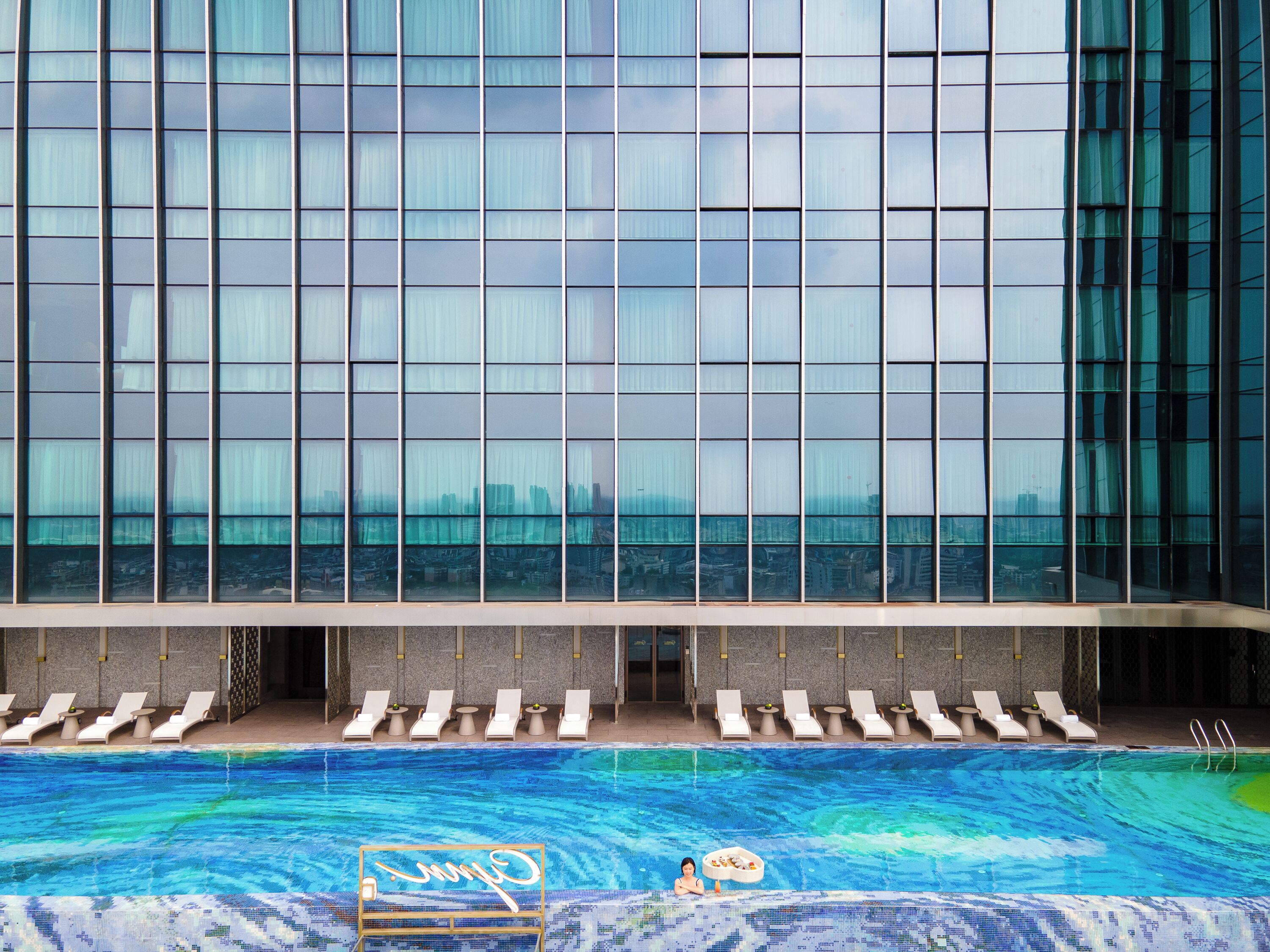Un hotel moderno con una amplia fachada de vidrio, un área de piscina con sillones de descanso y una vista despejada del horizonte de la ciudad.