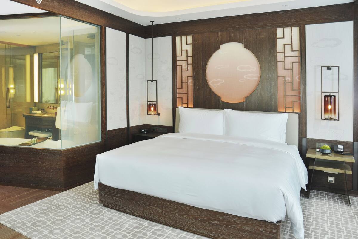 A modern hotel room with a large bed, a glass-enclosed shower area, and a decorative wall panel.