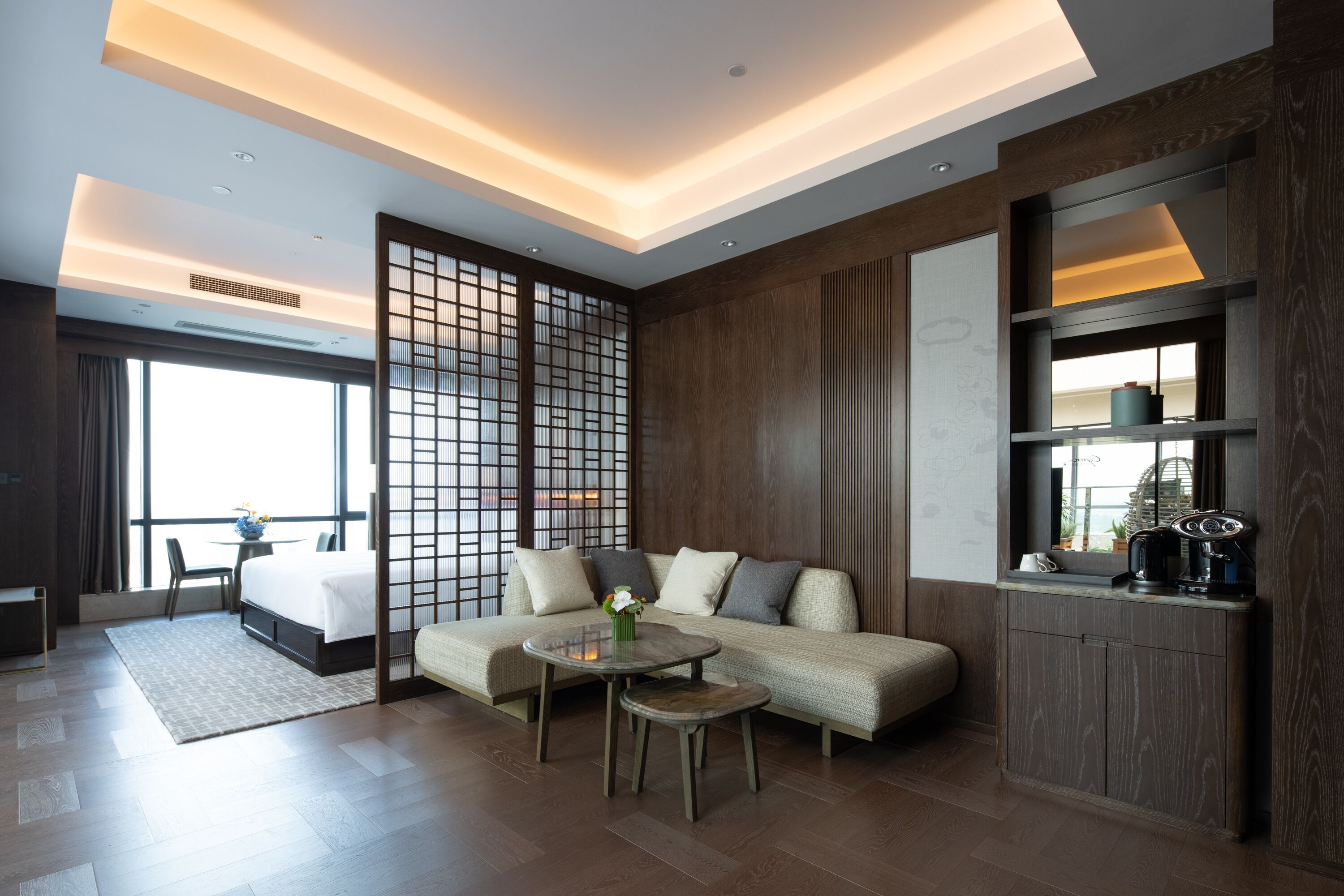 A modern hotel room with a sofa, a small round table, a bed, and a large window.