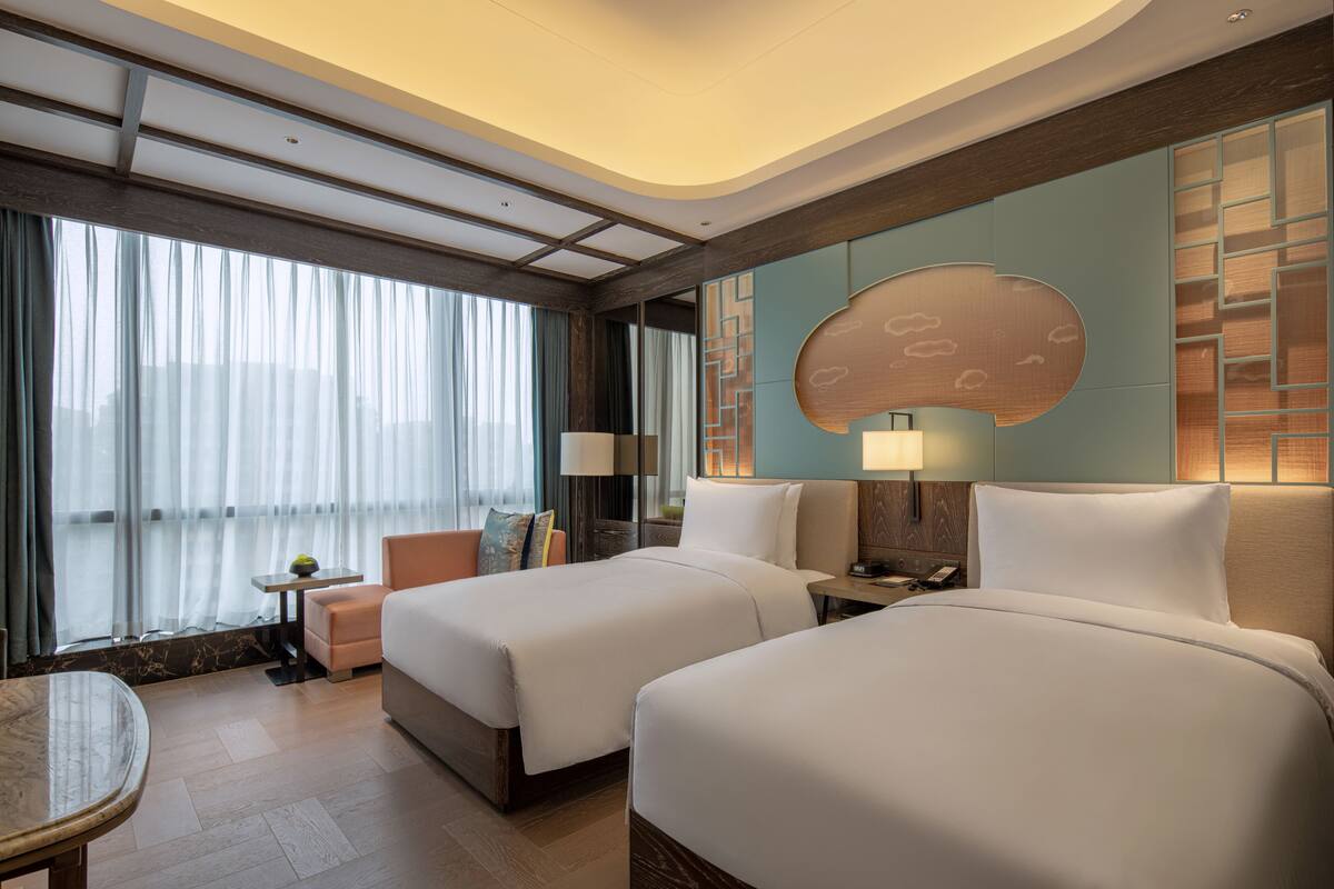 A hotel room with two beds, a small table, a sofa, and a large window with curtains.