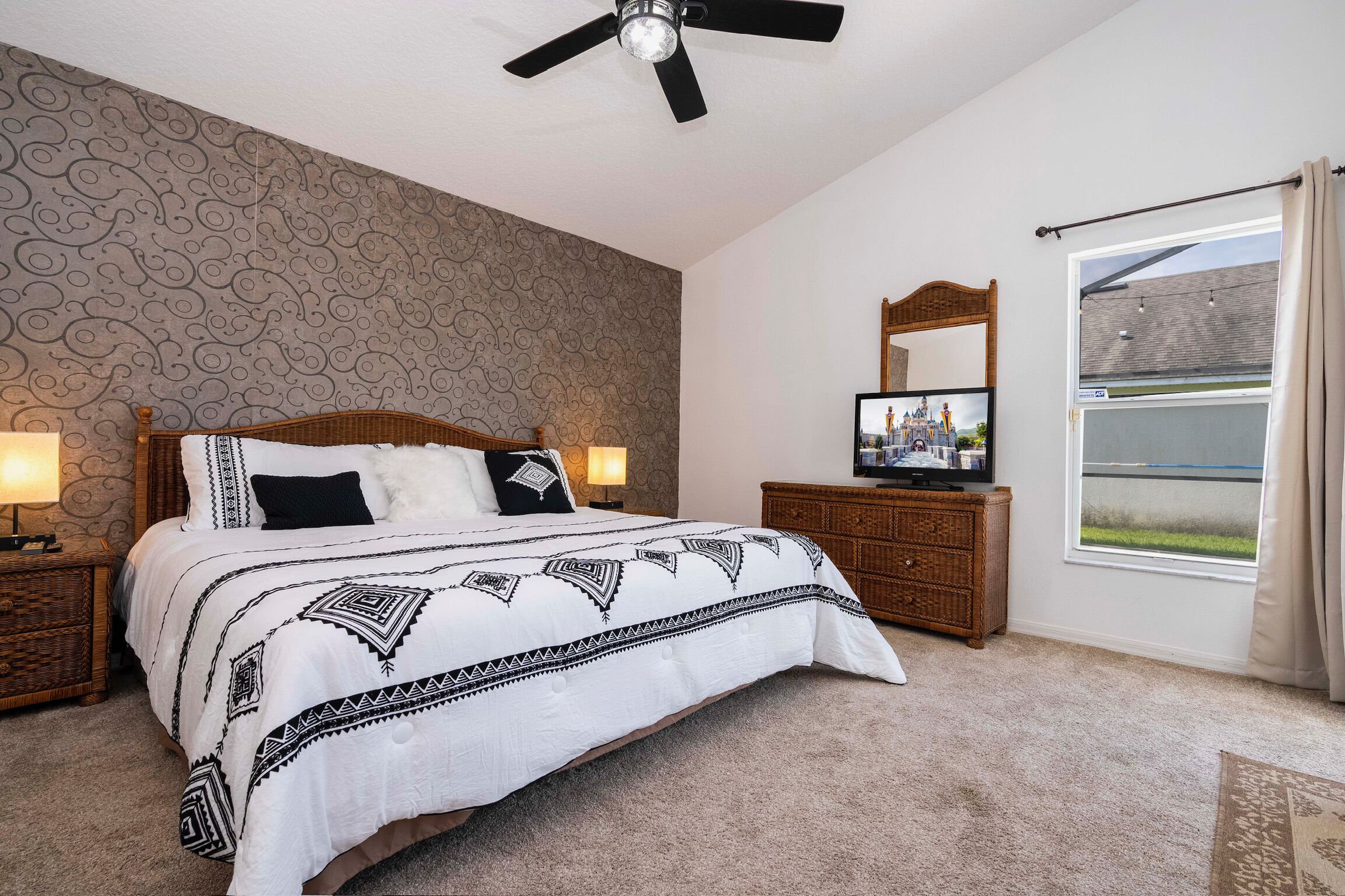 Une chambre à coucher avec un lit, un ventilateur de plafond, une télévision, une fenêtre avec des rideaux et un mur décoré d’un motif.