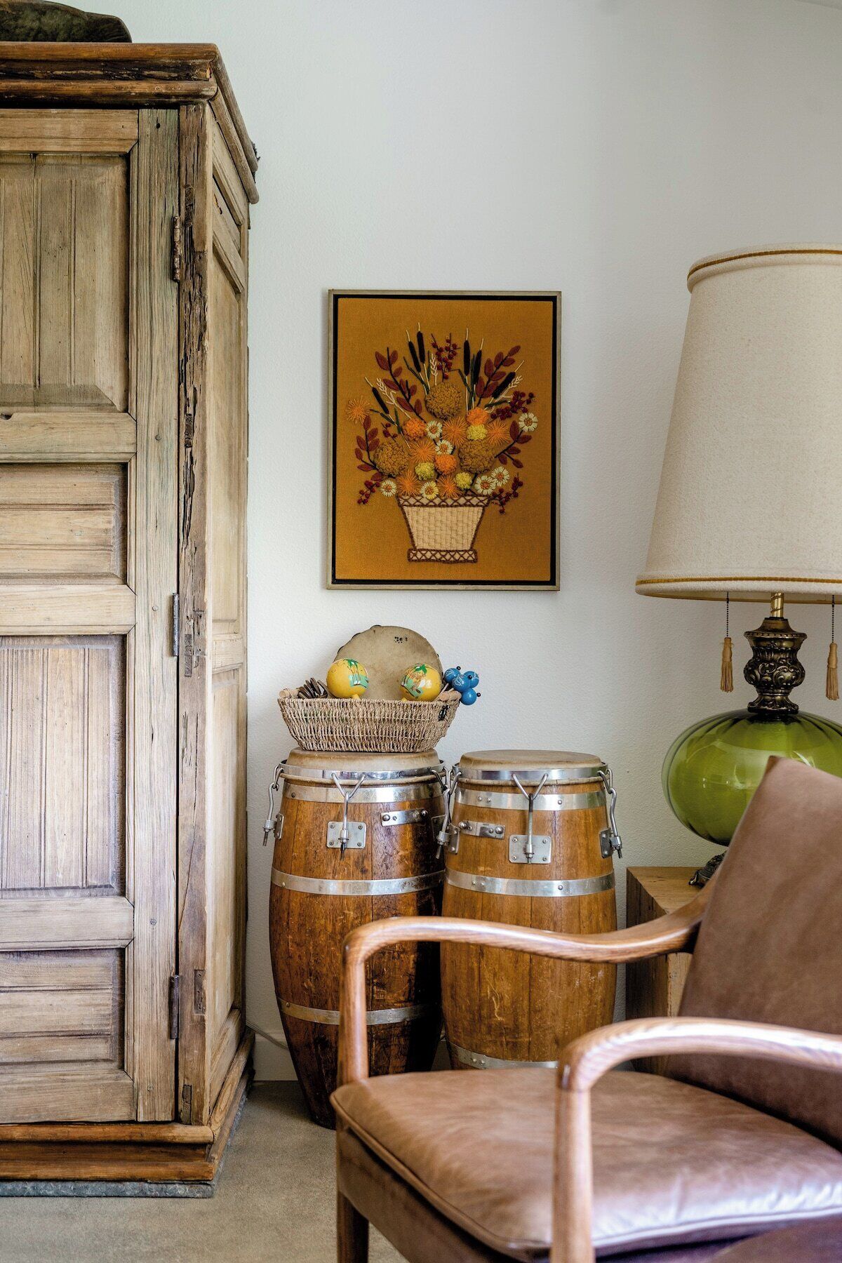 Una habitación rústica con un armario de madera, una silla, dos barriles de madera, una cesta de mimbre con frutas, un cuadro floral enmarco y una lámpara verde.