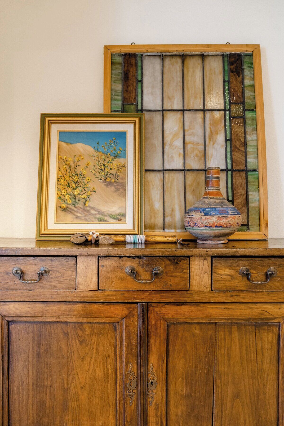 Um armário de madeira com gavetas, um quadro emoldurado de uma paisagem e uma janela de vitrais.