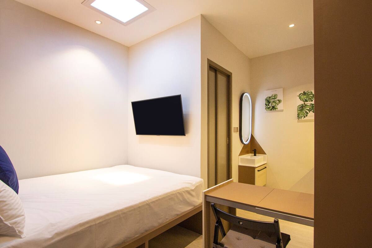 Una stanza piccola e ben illuminata con un letto singolo, una scrivania, uno specchio e una TV a parete.