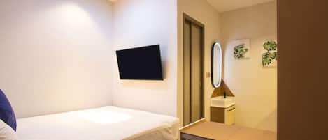 Una habitación pequeña y bien iluminada con una cama individual, un escritorio, un espejo y un televisor instalado en la pared.