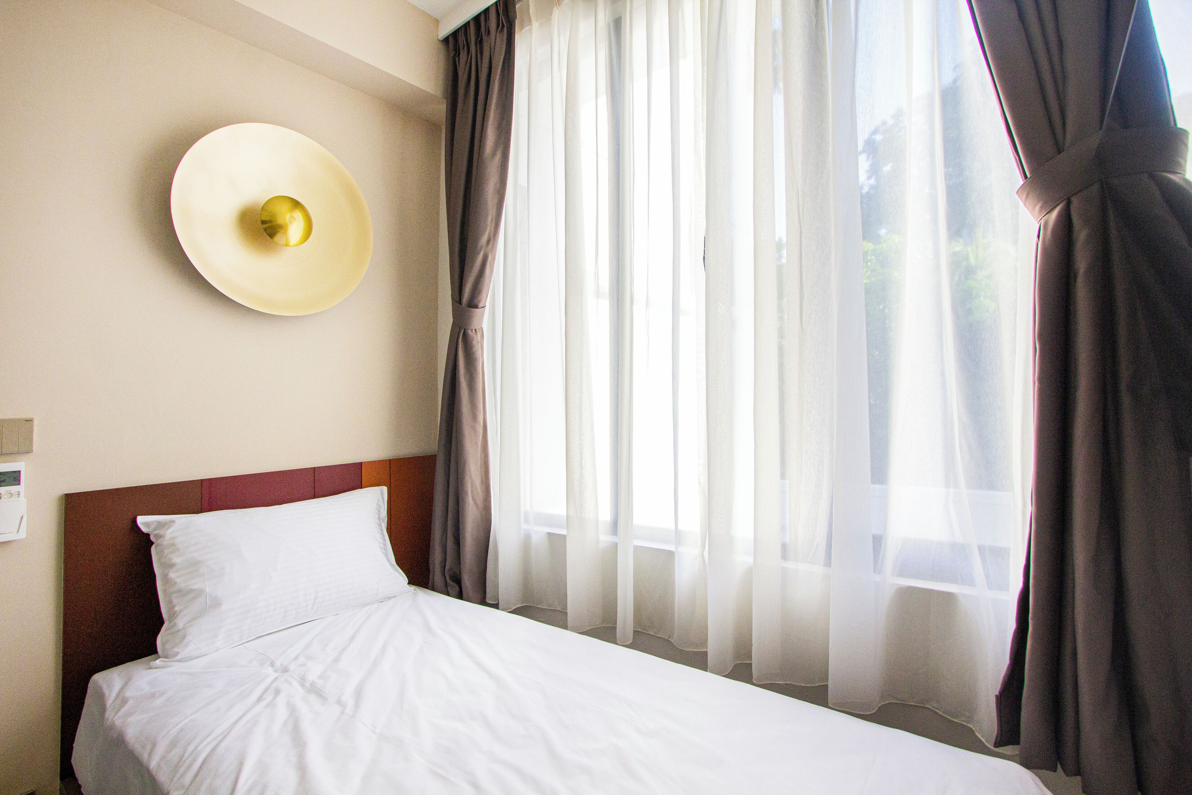 Una habitación de hotel con cama, lámpara de pared y cortinas traslúcidas.