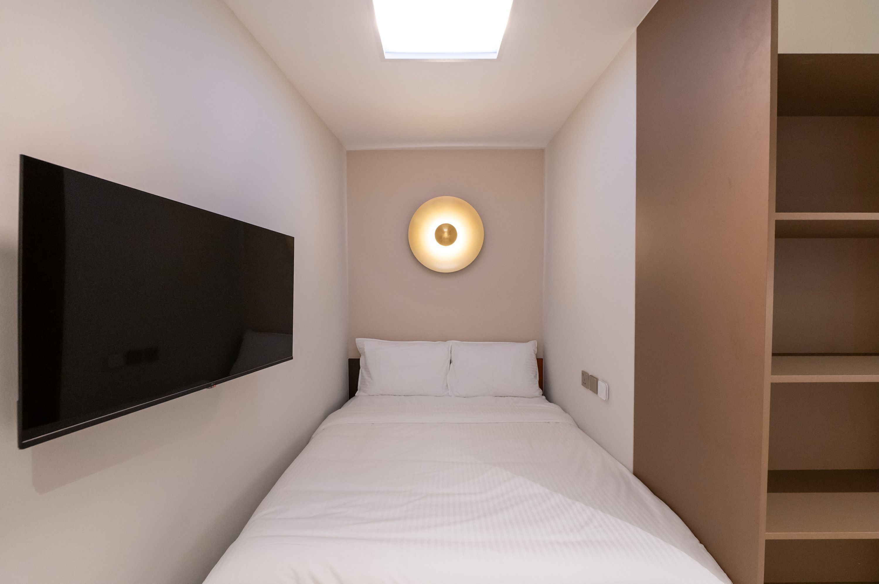 A narrow hotel room with a bed, a mounted TV, and a skylight.