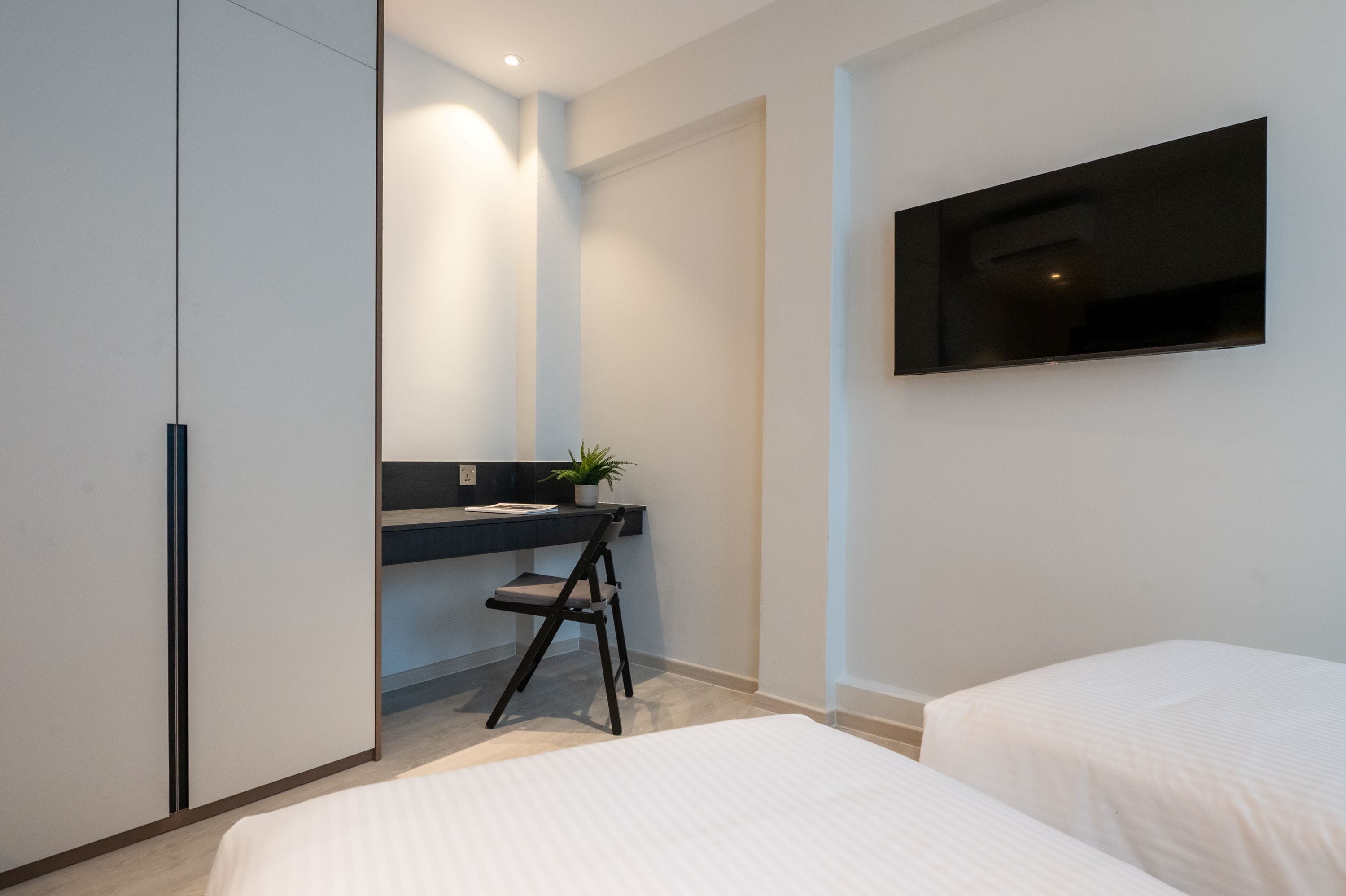 A modern bedroom with a bed, a desk with a chair, a potted plant, and a TV mounted on the wall.