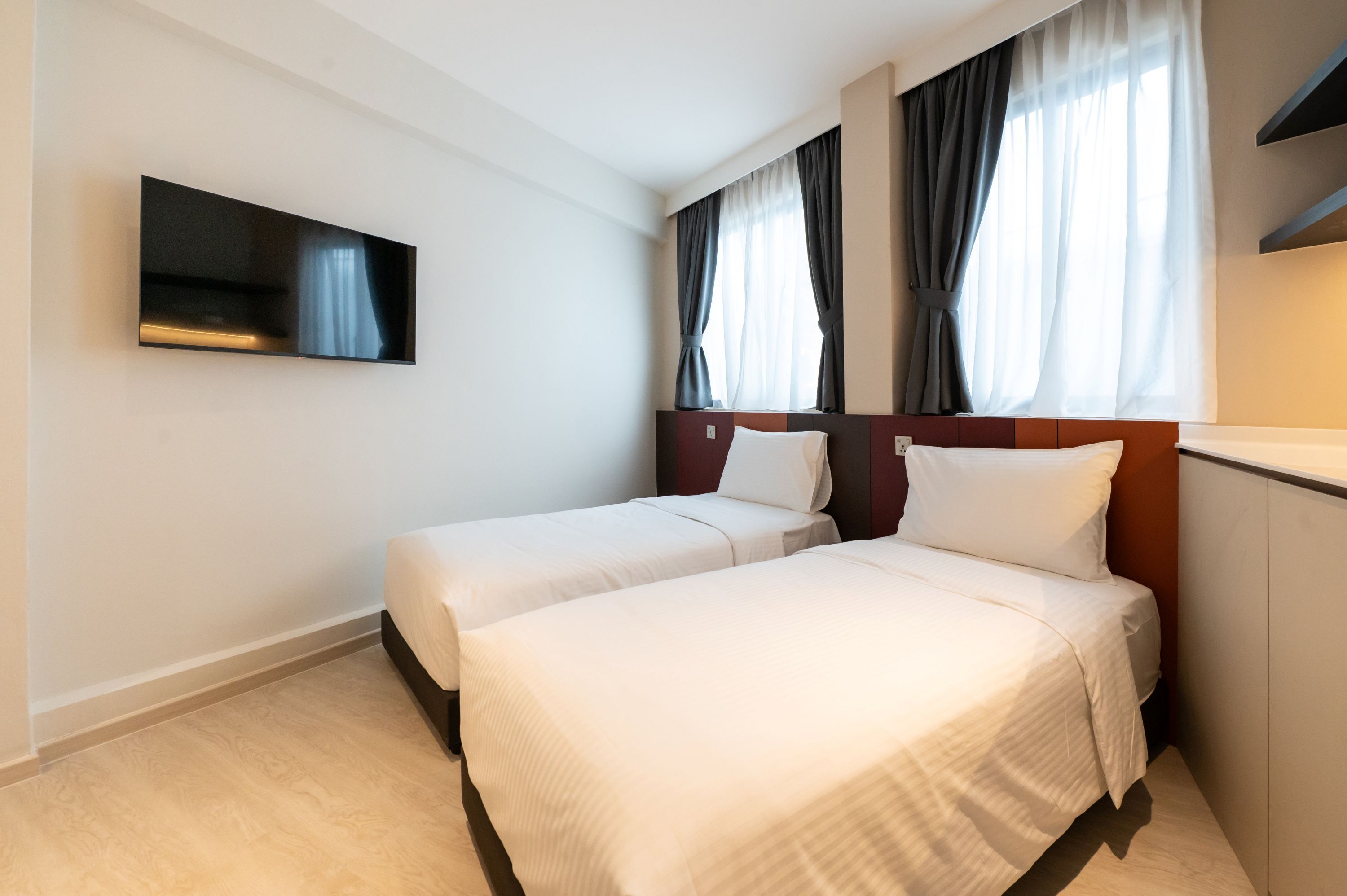 A hotel room with two single beds, a flat-screen TV mounted on the wall, and a large window with curtains.