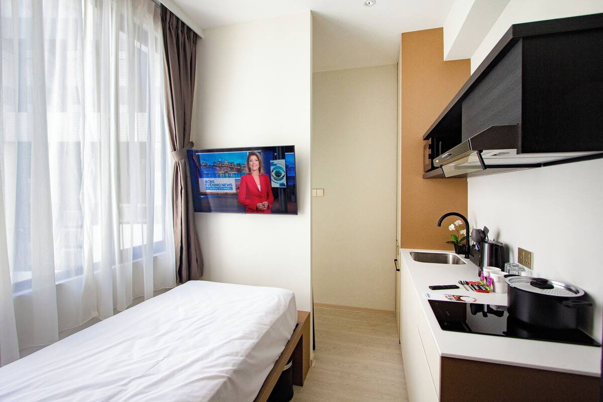 Una camera d'albergo con un letto, una TV che mostra un notiziario, un angolo cottura con lavandino e una finestra con tende leggere.