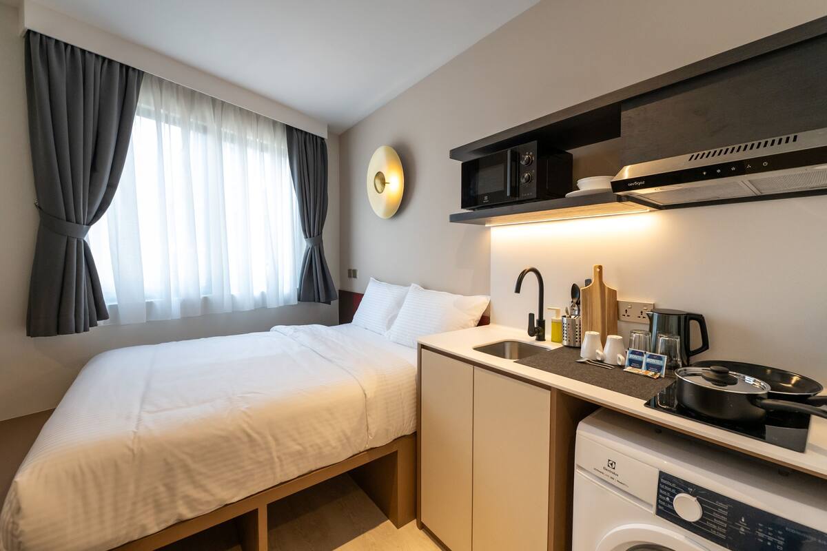 Una camera d'albergo compatta con un letto, un angolo cottura, una lavatrice e una finestra con tende.