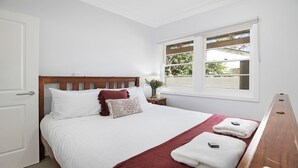 Een slaapkamer met een houten hoofdbord, wit beddengoed, een raam met zonwering en een nachtkastje met een lamp en een vaas.