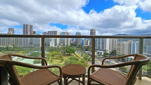 Un balcon agrémenté de chaises en osier et d’une petite table, d’où l’on peut admirer un paysage urbain avec ses immeubles de grande hauteur et un parc.