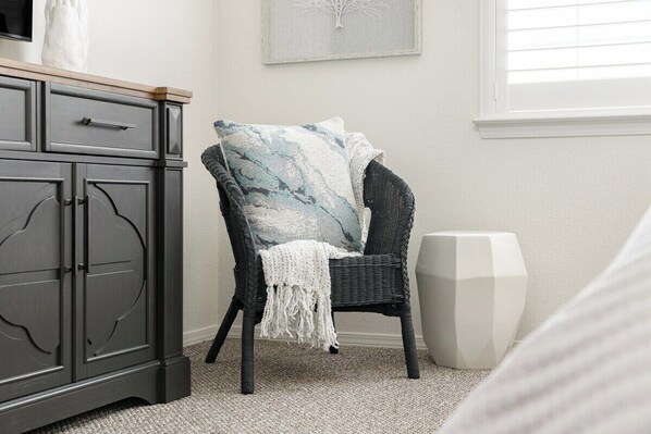 Una habitación con una silla de mimbre oscura, un cojín blanco, un armario y un altavoz blanco.