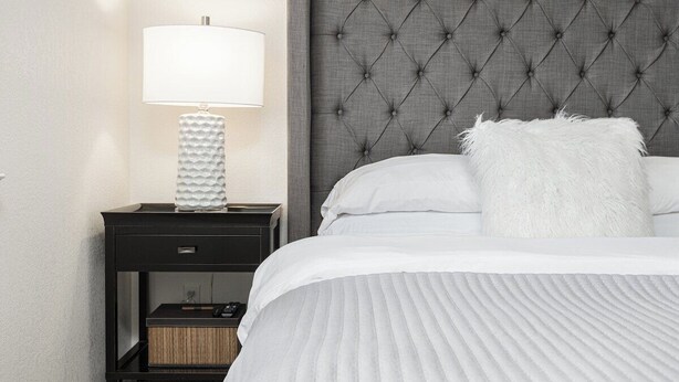 A neatly made bed with a tufted headboard, a bedside lamp, and a small nightstand with a drawer and a box.
